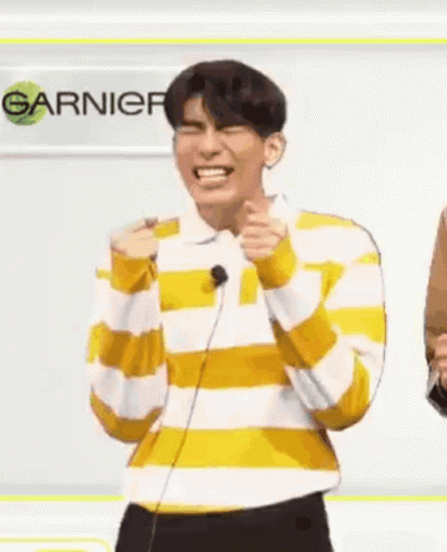 A Man In A Yellow And White Striped Shirt Is Laughing While Standing In Front Of A Microphone And A Sign . GIF
