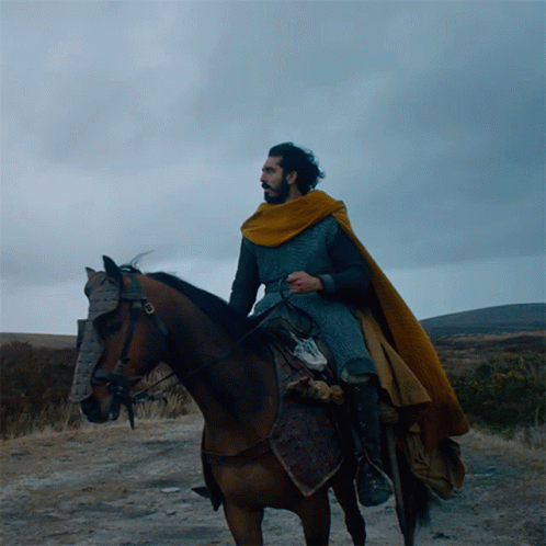 A Man In A Yellow Cape Is Riding A Horse On A Dirt Road GIF