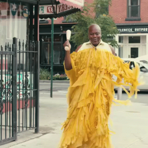 A Man In A Yellow Dress Is Walking Down The Street In Front Of A Store Called Ttleneck Outpost GIF