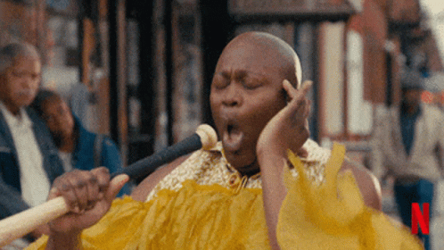 A Man In A Yellow Dress Singing Into A Microphone With The Letter N In The Corner GIF