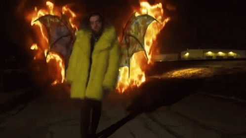 A Man In A Yellow Fur Coat Is Standing In Front Of Flames . GIF