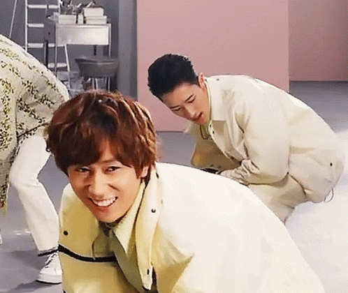 A Man In A Yellow Jacket Is Smiling While Another Man Squats Behind Him GIF