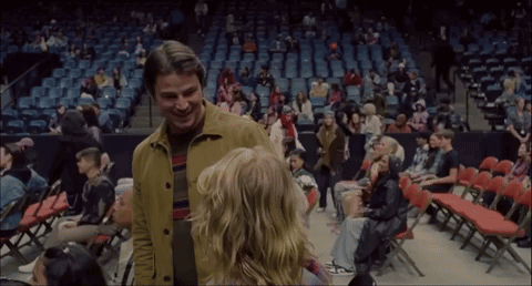 A Man In A Yellow Jacket Stands Next To A Woman In A Stadium GIF