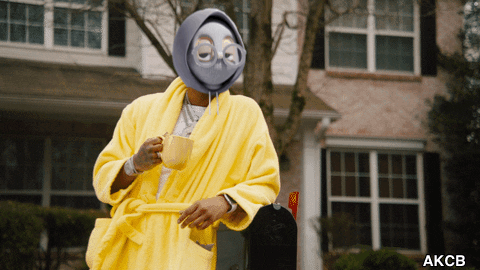 A Man In A Yellow Robe Is Holding A Cup Of Coffee In Front Of A House GIF
