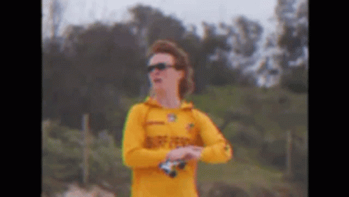 A Man In A Yellow Shirt And Sunglasses Is Standing On A Beach Holding Binoculars . GIF