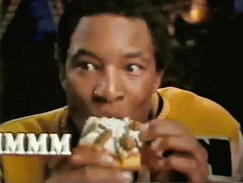 A Man In A Yellow Shirt Is Eating A Hamburger With The Word Mmmm In The Background GIF