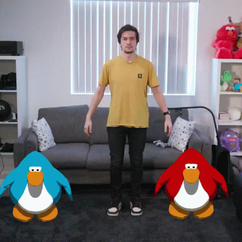A Man In A Yellow Shirt Stands In Front Of A Couch And Two Penguins GIF
