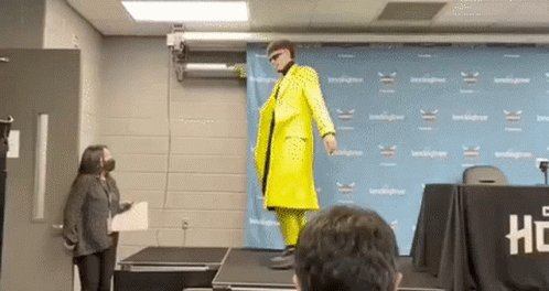 A Man In A Yellow Suit Is Standing On A Stage In Front Of A Crowd . GIF