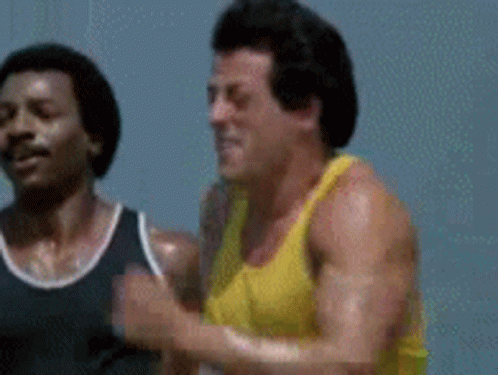 A Man In A Yellow Tank Top Is Running Next To Another Man . GIF