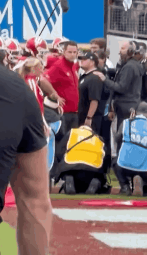 A Man In A Yellow Vest Is Kneeling On A Football Field With A Group Of People Standing Around Him . GIF
