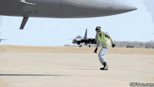 A Man In A Yellow Vest Is Standing In Front Of An Airplane With A Gifsec.com Watermark GIF