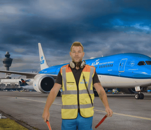 A Man In A Yellow Vest Stands In Front Of A Blue Klm Plane GIF