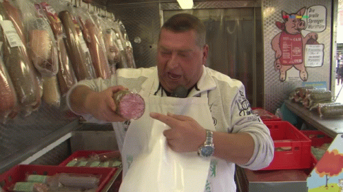 A Man In An Apron Holds A Piece Of Meat In Front Of A Sign That Says " Die Größte Europäische Wurst " GIF