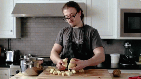 A Man In An Apron Is Peeling Potatoes On A Wooden Cutting Board GIF