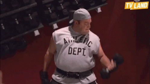 A Man In An Athletic Dept Shirt Is Lifting Dumbbells In A Gym GIF