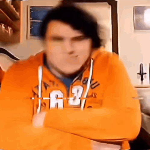A Man In An Orange Hoodie Is Standing With His Arms Crossed . GIF