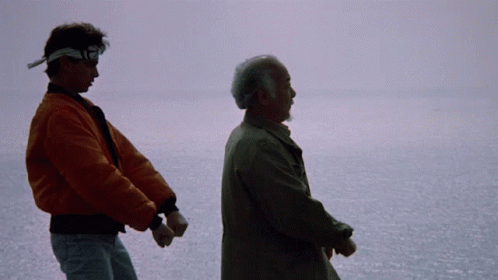 A Man In An Orange Jacket Holds The Hand Of An Older Man In A Brown Coat GIF