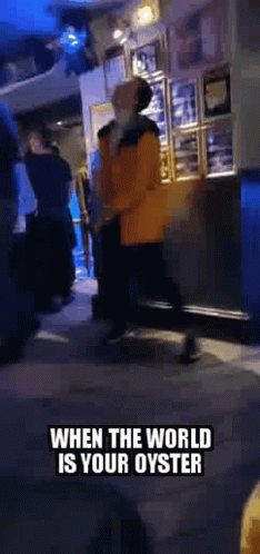A Man In An Orange Jacket Is Walking Down A Street With The Words When The World Is Your Oyster On The Bottom . GIF