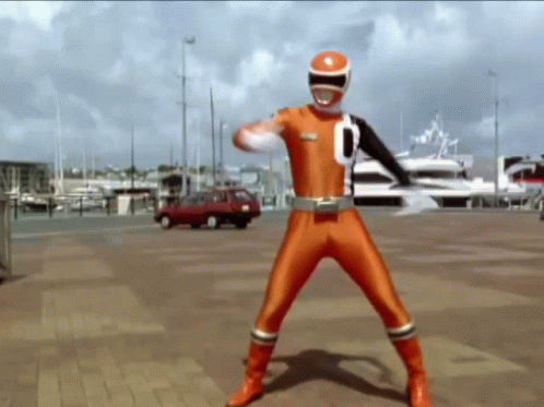 A Man In An Orange Power Ranger Costume Is Standing On A Brick Sidewalk In Front Of A Boat . GIF