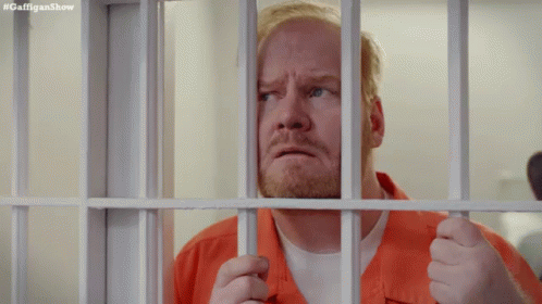 A Man In An Orange Shirt Is Behind Bars In A Prison Cell . GIF