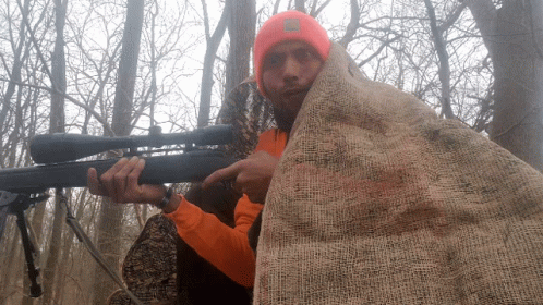 A Man In An Orange Shirt Is Holding A Rifle And Pointing GIF