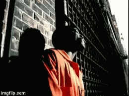 A Man In An Orange Shirt Is Standing In Front Of A Brick Wall . GIF