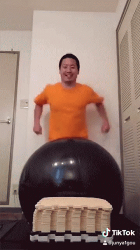 A Man In An Orange Shirt Is Standing On A Large Black Ball With A Tiktok Watermark GIF