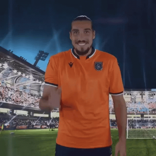 A Man In An Orange Shirt Is Standing On A Soccer Field With His Arms Outstretched GIF