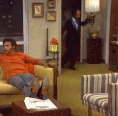 A Man In An Orange Shirt Sits On A Couch In A Living Room GIF