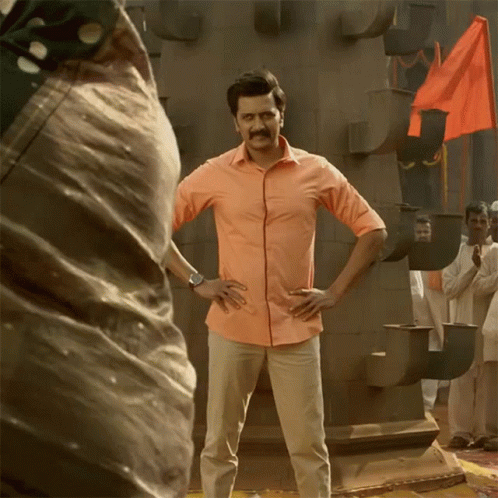 A Man In An Orange Shirt Stands With His Hands On His Hips In Front Of A Crowd GIF