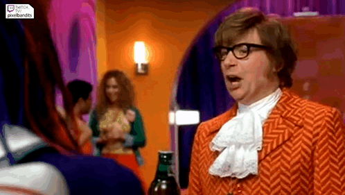 A Man In An Orange Suit And Glasses Is Standing In A Room Talking To A Woman . GIF