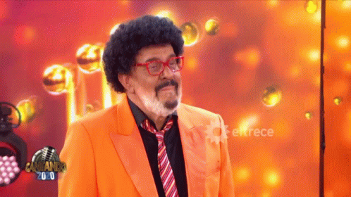 A Man In An Orange Suit And Tie Is Standing In Front Of A Sign That Says Cantando 2.0 GIF