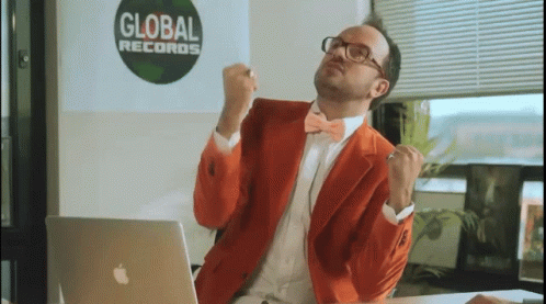 A Man In An Orange Suit Is Sitting In Front Of An Apple Laptop In Front Of A Global Records Sign GIF