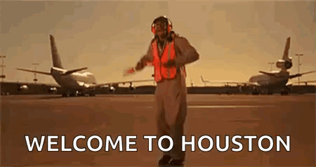 A Man In An Orange Vest Is Standing On A Runway In Front Of Planes . GIF