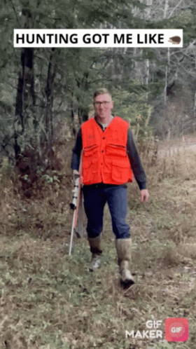 A Man In An Orange Vest Is Walking Through The Woods With A Rifle GIF