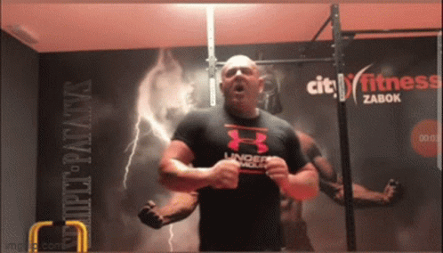 A Man In An Under Armour Shirt Is Standing In Front Of A City Fitness Zabok Sign GIF