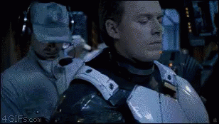 A Man In Armor Is Standing Next To A Man Wearing A Hat And Headphones . GIF