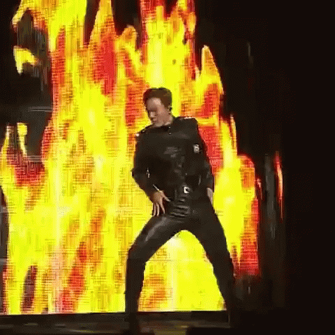 A Man In Leather Pants Is Dancing In Front Of A Large Screen With Flames On It GIF