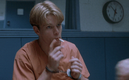 A Man In Orange Scrubs Is Sitting In Front Of A Wall Clock That Says Sounds Like GIF