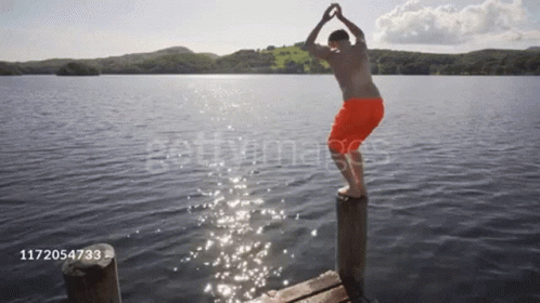 A Man In Orange Shorts Is Jumping Into A Lake From A Pier GIF
