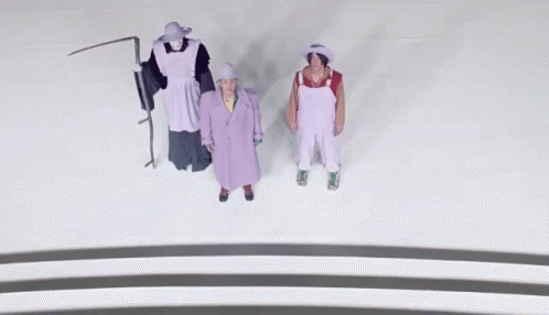 A Man In Overalls And A Woman In A Purple Coat Are Standing Next To Each Other GIF
