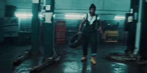 A Man In Overalls Is Holding A Tire In A Dark Garage . GIF
