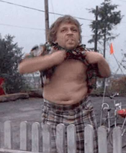 A Man In Plaid Shorts Is Standing In Front Of A Fence And Taking Off His Shirt GIF