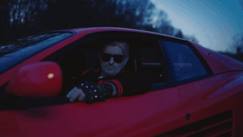 A Man In Sunglasses Is Driving A Red Car GIF