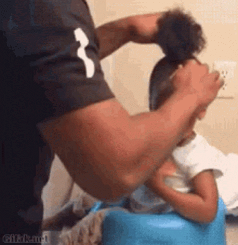 A Man Is Brushing A Little Girl 'S Hair While She Is Sitting On A Blue Chair . GIF