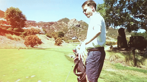 A Man Is Carrying A Golf Bag And A Golf Club On A Golf Course GIF