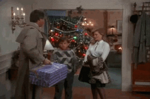 A Man Is Carrying A Purple Gift Box In Front Of A Christmas Tree GIF