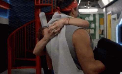 A Man Is Carrying A Woman In His Arms In A Room With A Spiral Staircase GIF