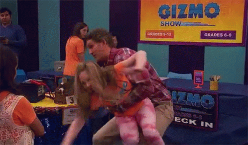 A Man Is Carrying A Woman In His Arms In Front Of A Sign That Says Gizmo Show . GIF