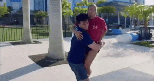 A Man Is Carrying A Woman On His Back In A Park While A Woman Looks On . GIF